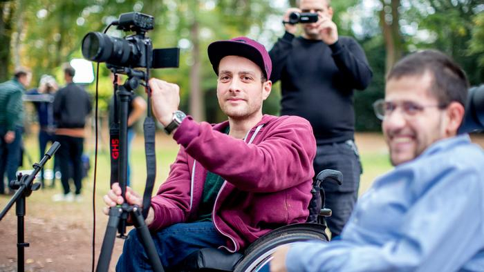 Zwei junge Männer im Rollstuhl bedienen einen Kamera auf einem Stativ.
