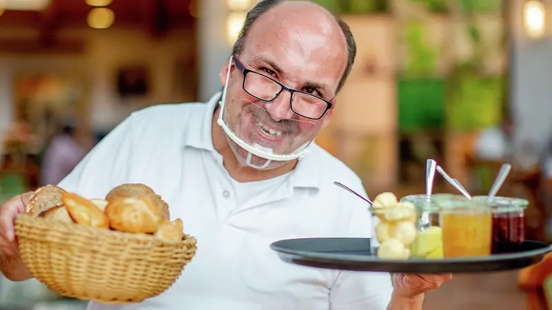 Ein Mann mit durchsichtigem Mundschutz halt ein Tablet mit Marmelade und ein Korb mit Brötchen in den Händen.