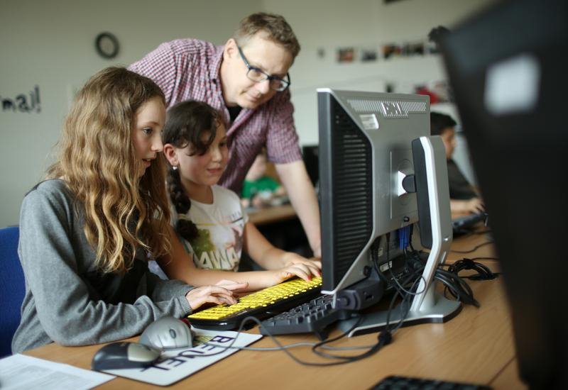 Zwei Mädchen sitzen vor einem Computerbildschirm und bedienen die Tastatur. Hinter ihnen steht ein Lehrer und schaut ihnen zu. 