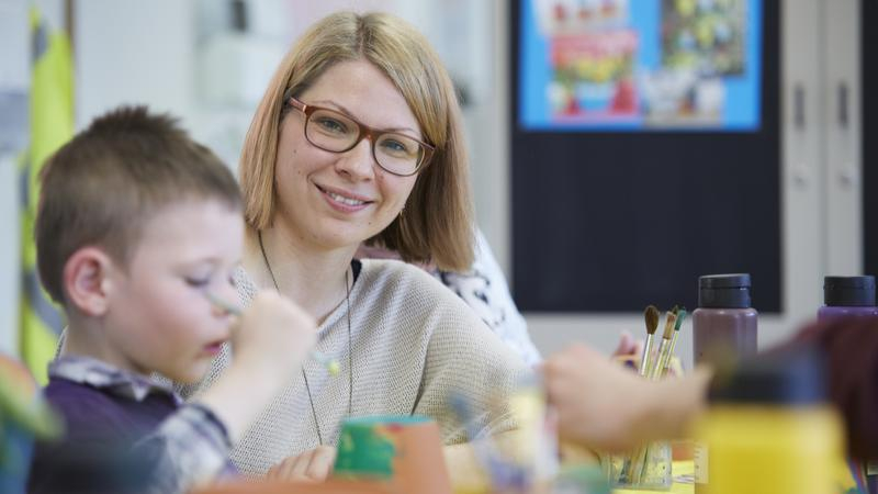 Eine blonde Frau sitzt mit einem kleinen Jungen an einem Tisch. Darauf stehen Farbflaschen und Gläser mit Pinseln. 