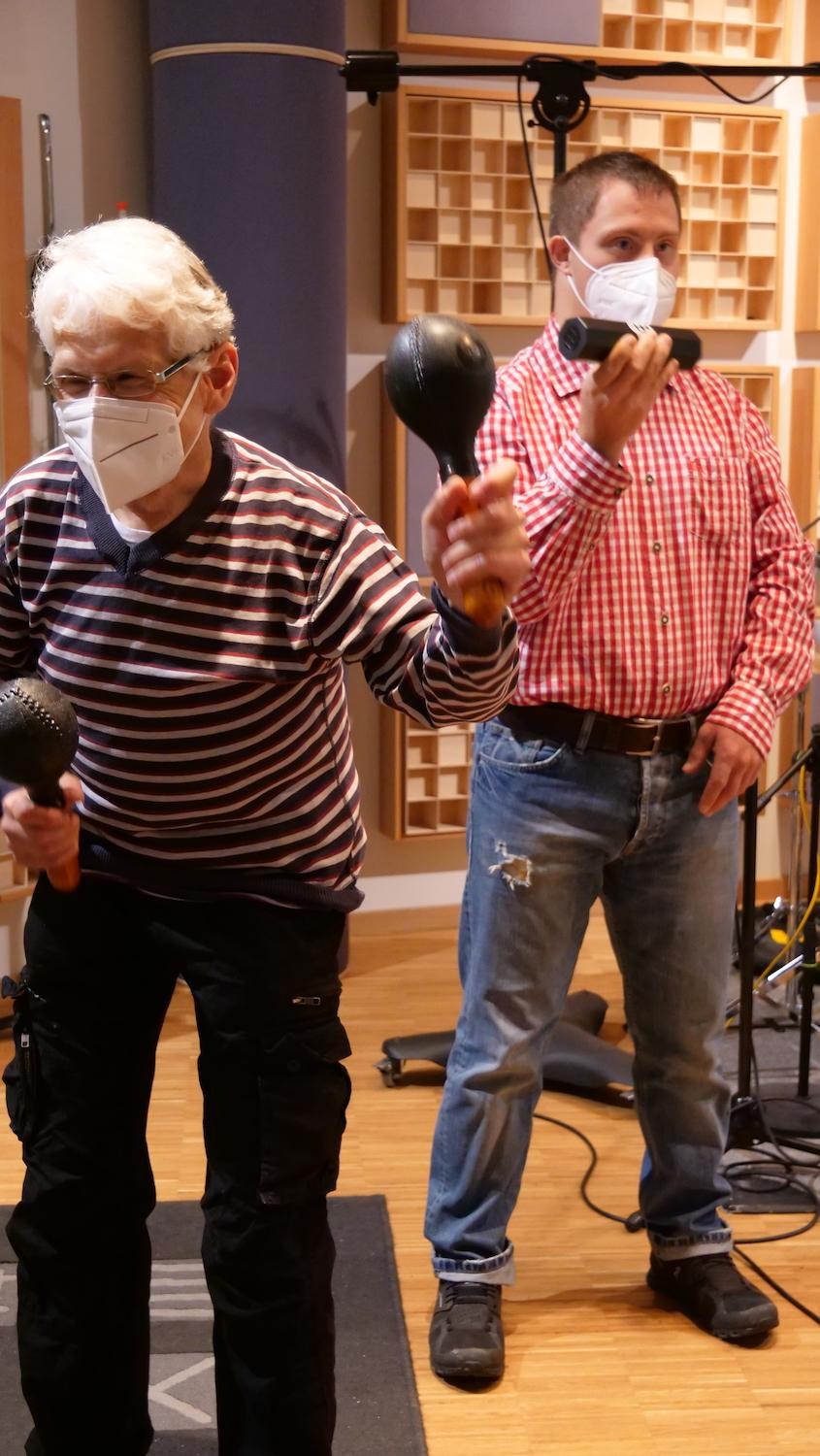 Eine älterer und ein jüngerer Mann stehen im Tonstudio und bewegen jeweils eine Rassel.