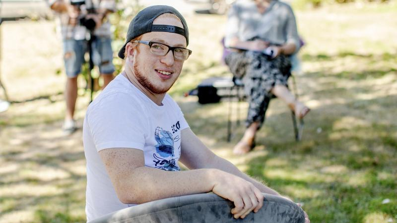 Ein junger Mann mit Brille sitzt auf eine Stuhl. Er dreht sich lächelnd zur Kamera. Im Hintergrund stehen weitere Personen mit Kameras.