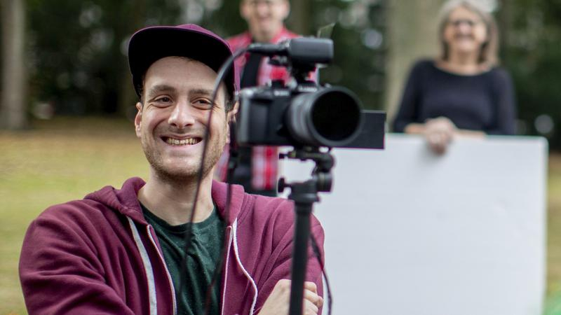 Ein Mann sitzt hinter einer Videokamera und filmt eine Person außerhalb des Bildes.