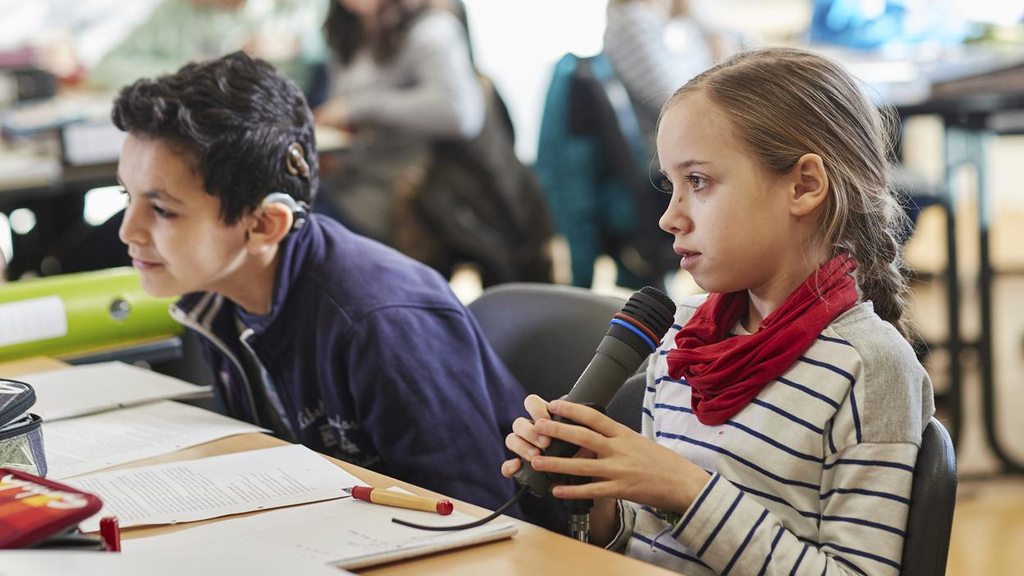 Ein Junge und ein Mädchen sitzen in einer Klasse. Das Mädchen spricht in ein Mikrofon. 
