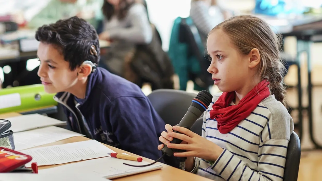 Ein Junge und ein Mädchen sitzen in einer Klasse. Das Mädchen spricht in ein Mikrofon. 