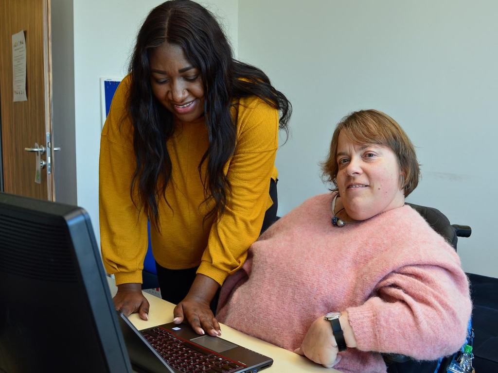 Eine Frau im Rollstuhl sitzt im Büro vor dem PC, neben ihr steht eine junge schwarze Frau