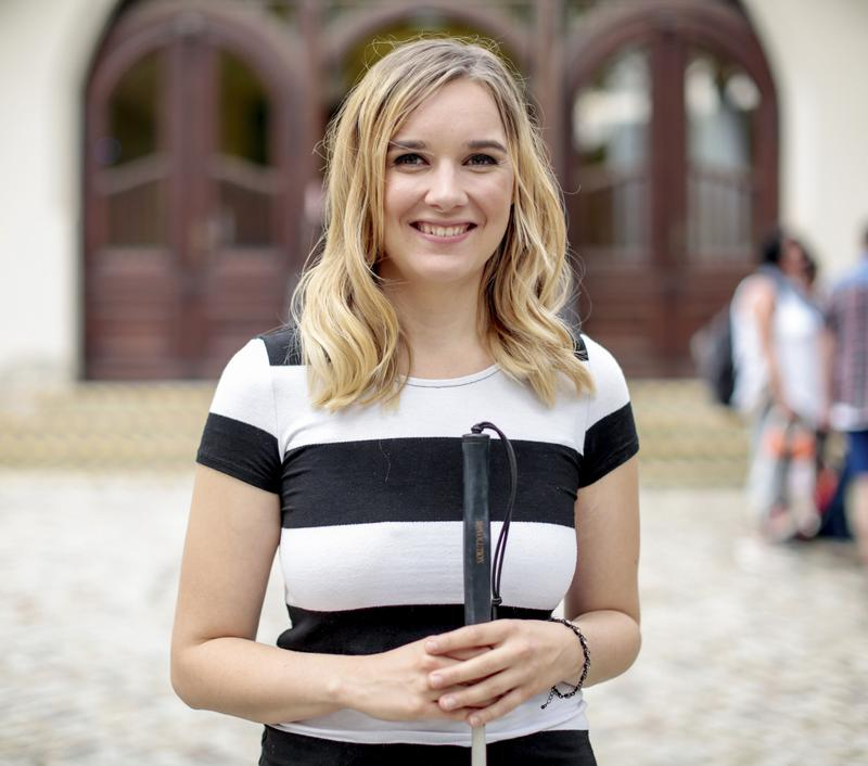 Eine junge Frau in  gestreiften T-Shirt lächelt in die Kamera. In den Händen hält sie vor sich einen Langstock.