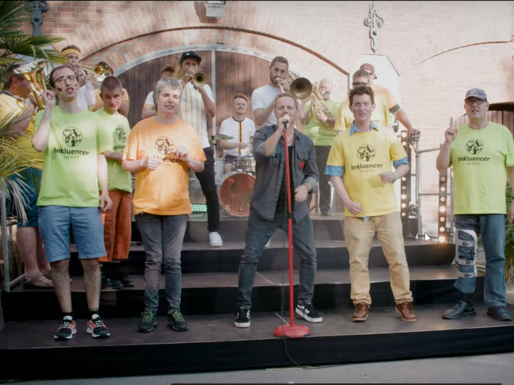 Sänger*innen mit und ohne Behinderung stehen gemeinsam mit einer Brass-Band auf der Bühne und singen.