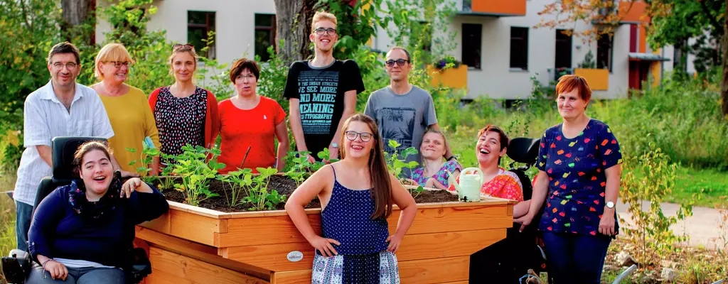 Eine Gruppe von Menschen mit und ohne Behinderung steht um ein Hochbeet, das sie gerade bepflanzt haben. Alle lächeln.