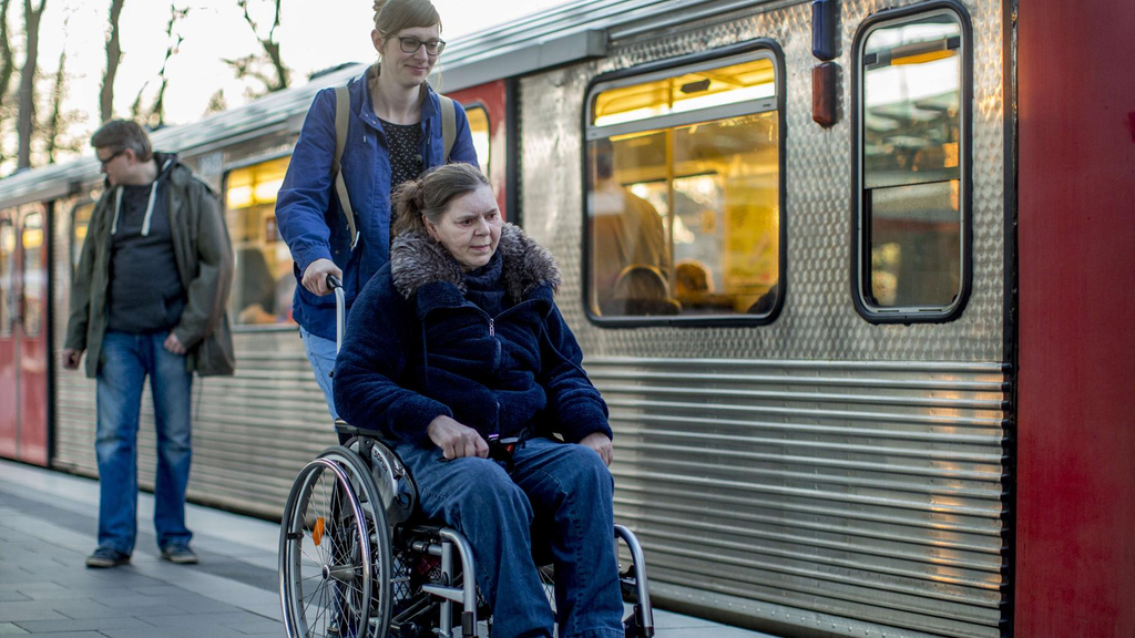 Eine ältere Frau sitzt in einem Rollstuhl, der von einer jüngeren Frau geschoben wird. Die beiden sind an einem Bahnsteig, an dem eine Bahn steht.