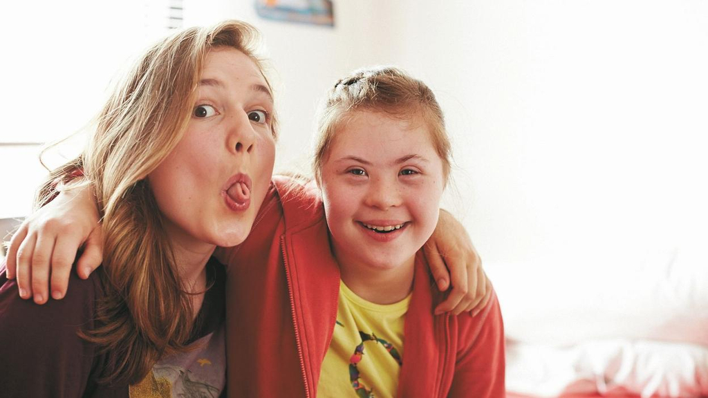 Zwei jugendliche Freundinnen mit und ohne Down-Syndrom Arm in Arm. Eine lacht in die Kamera, die zweite zieht eine lustige Grimasse.