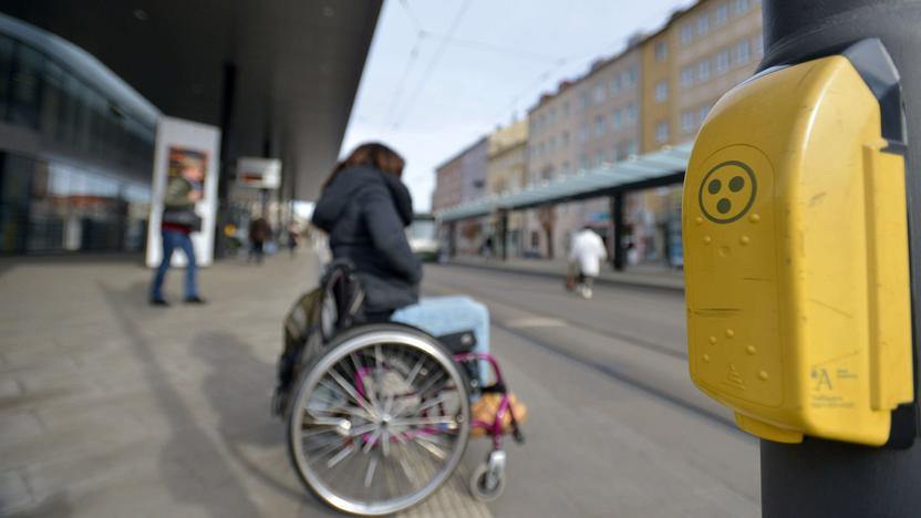 Eine Frau um Rollstuhl an einer barrierefreien Haltestelle, im Vordergrund eine Blindenampel