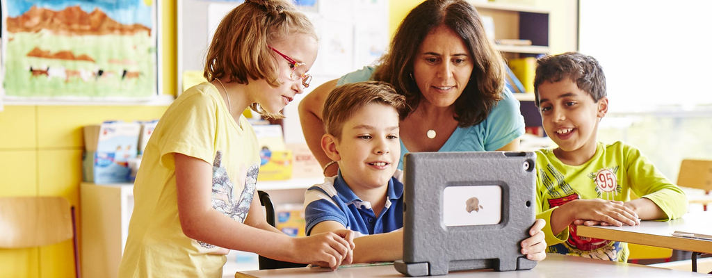 Zwei Jungen und ein Mädchen schauen in einem Klassenzimmer gemeinsam auf ein Tablet. Hinter ihnen steht die Lehrerin. Einer der Jungen sitzt im Rollstuhl.
