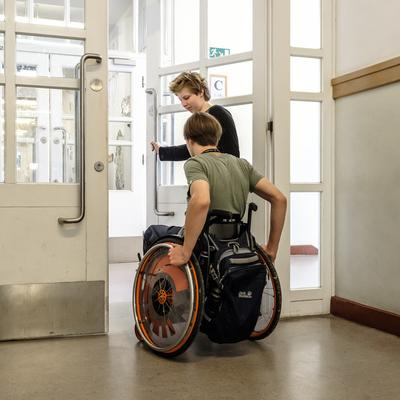 Eine Schülerin hält einem Schüler im Rollstuhl eine Tür im Flur auf 