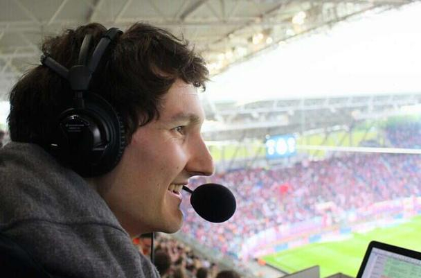 Porträt eines jungen Mannes mit Kopfhörer und Mikro bei der Audiodeskription in einem Stadion.