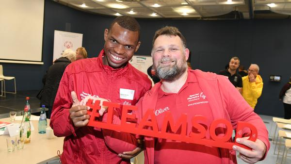 Ein junger schwarzer Mann und ein weißer Mann mittleren Alters stehen lächelnd nebeneinander und halten den Schriftzug #TEAMSOD fest