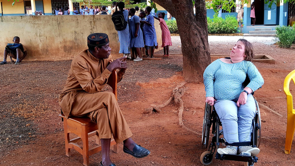 Eine Frau im Rollstuhl und ein schwarzer Mann in traditioneller Kleidung im Gespräch unter einem Baum vor einer afrikanischen Schule.