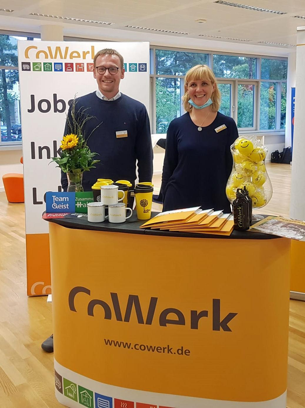 Ein Mann und eine Frau stehen in einem Konferenzsaal hinter einem Stand, auf dem CoWerk steht