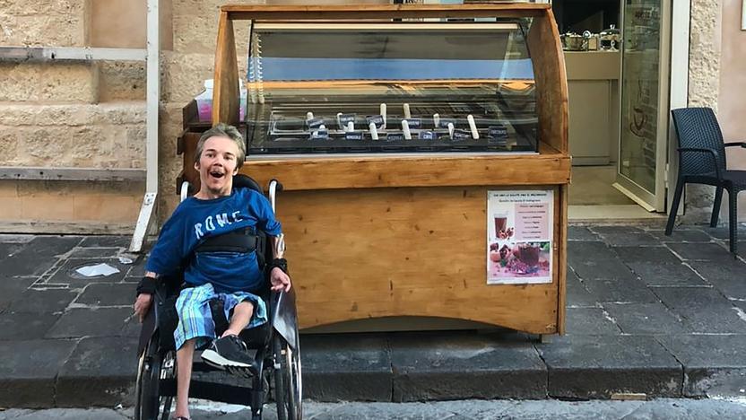 Ein Mann sitzt in einem Rollstuhl in einer Fußgängerzone. Hinter ihm steht vor einer Eisdiele eine Eistheke. 