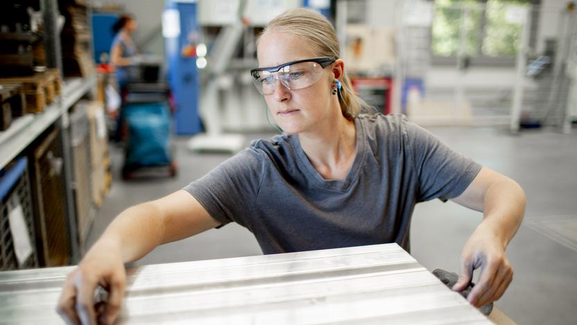 Eine junge Frau mit blonden Haaren steht in einer großen Werkstatt. Sie trägt eine Schutzbrille und bearbeitet ein Metallblech.