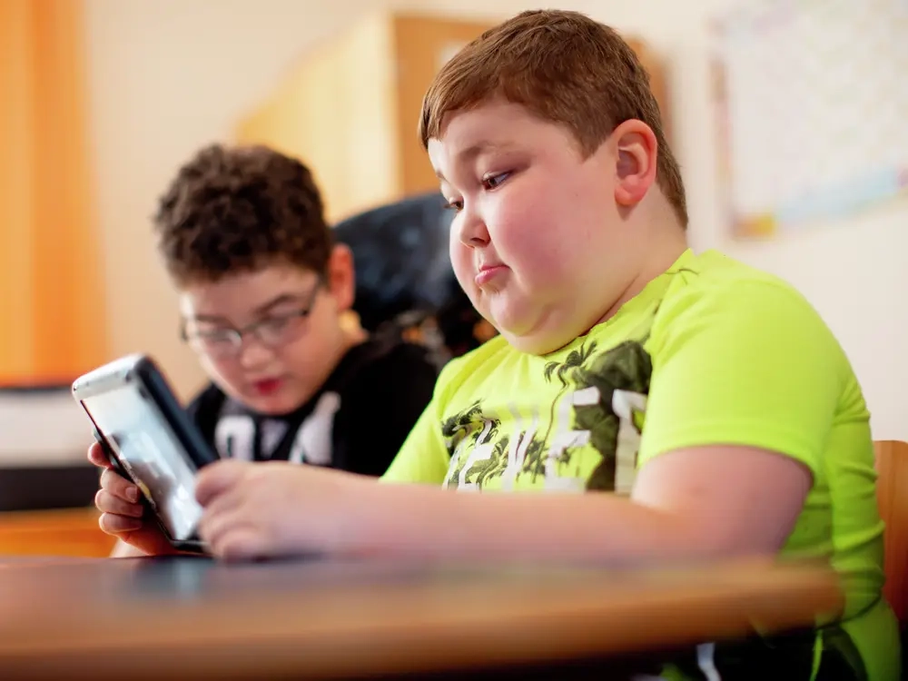 Zwei Jungen sitzen nebeneinander in einem Klassenzimmer. Der Junge im Vordergrund hält ein Tablet in den Händen.