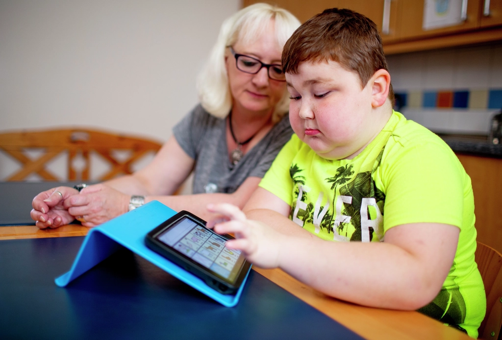 Ein Junge in einem grellgelben T-Shirt sitzt neben einer blonden Frau und bedient ein Tablet. 