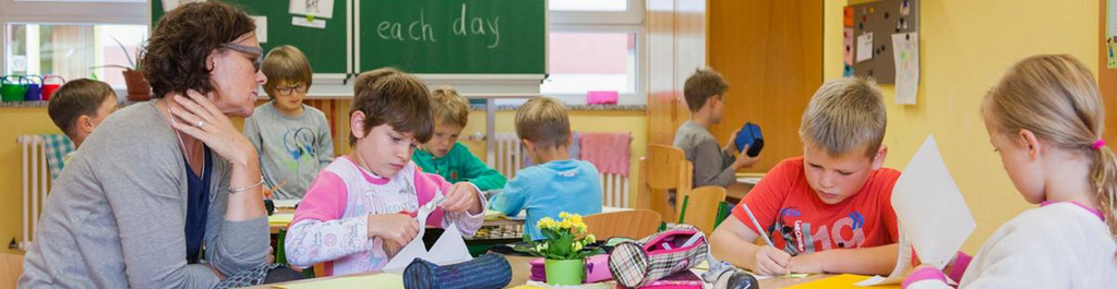 Lehrerin und drei Kinder an einem Tisch in einem Klassenzimmer. Im Hintergrund sieht man eine Tafel.
