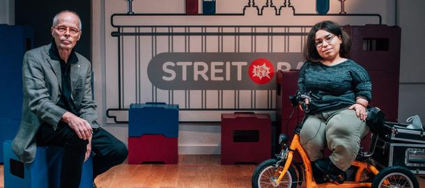 Ein Mann in höherem Erwachsenenalter und eine junge, kleinwüchsige Frau sitzen nebeneinander in einem Studio