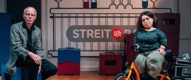 Ein Mann in höherem Erwachsenenalter und eine junge, kleinwüchsige Frau sitzen nebeneinander in einem Studio