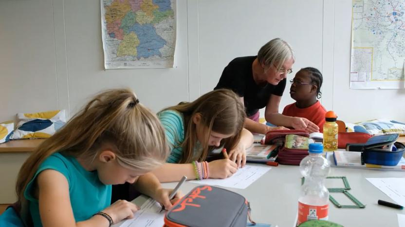 Eine Frau steht in einem Klassenzimmer an einem Tisch mit mehreren Schüler*innen und bespricht mit einem Kind eine Aufgabe. 