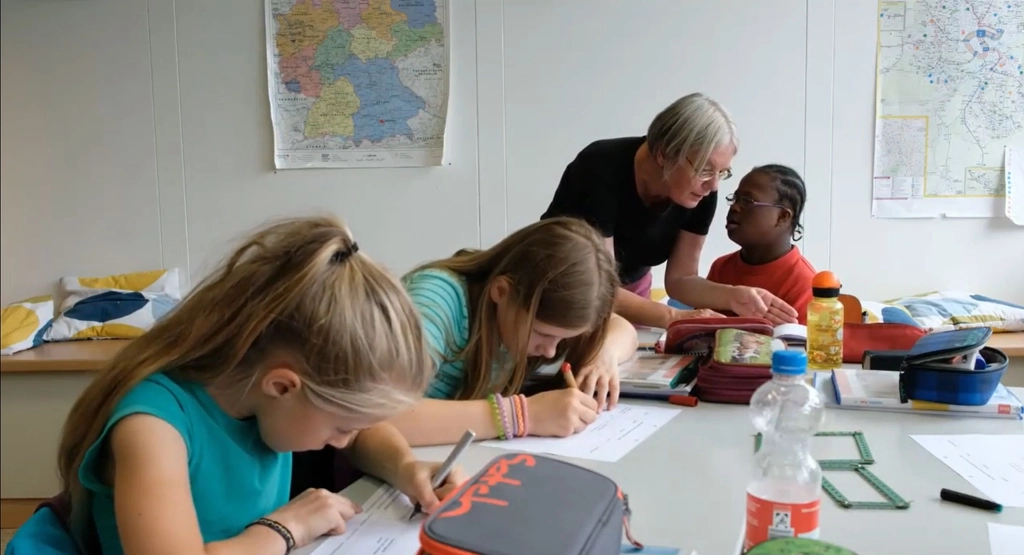 Eine Frau steht in einem Klassenzimmer an einem Tisch mit mehreren Schüler*innen und bespricht mit einem Kind eine Aufgabe. 