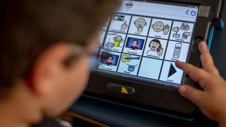 Ein Junge schaut auf ein Tablet. Dort sind verschiedene Piktogramme mit Erläuterung. Zum Beispiel "leiser", "lauter", "Informationen"