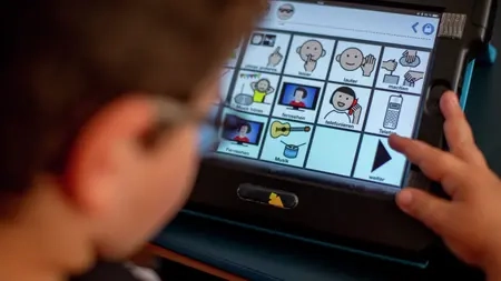 Ein Junge schaut auf ein Tablet. Dort sind verschiedene Piktogramme mit Erläuterung. Zum Beispiel "leiser", "lauter", "Informationen"