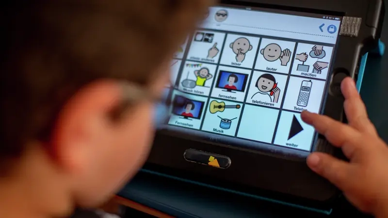 Ein Junge schaut auf ein Tablet. Dort sind verschiedene Piktogramme mit Erläuterung. Zum Beispiel "leiser", "lauter", "Informationen"