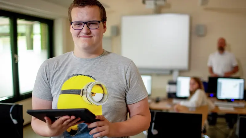 Eine Junge hält ein Tablet in der Hand und lacht in die Kamera. Im Hintergrund ist unscharf ein Klassenraum mit Computern zu sehen.