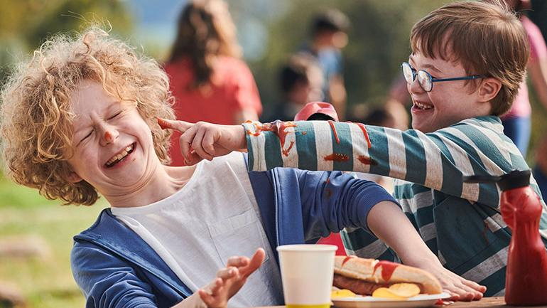 Zwei Jungen mit und ohne Behinderung sitzen an einem Tisch im Freien und spielen mit Ketchup.