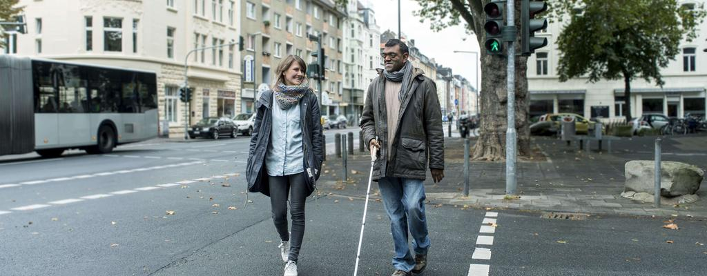 Eine Frau und ein Mann mit Langstock gehen über eine Ampel.