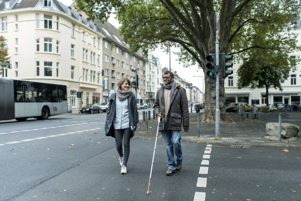 Eine Frau und ein Mann mit Langstock gehen über eine Ampel.