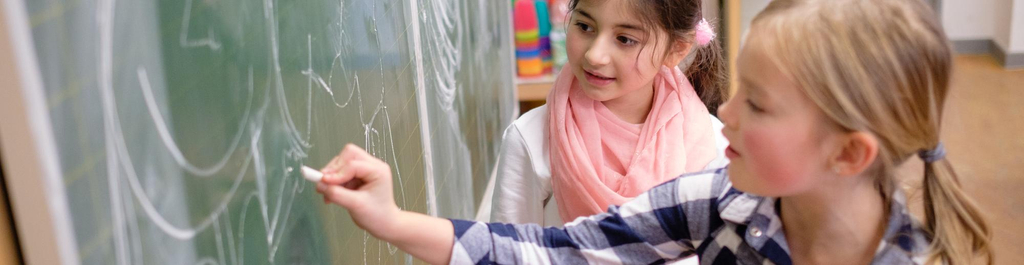 Ein Mädchen schreibt im Klassenraum an eine Tafel, ein anderes sieht ihr dabei zu.