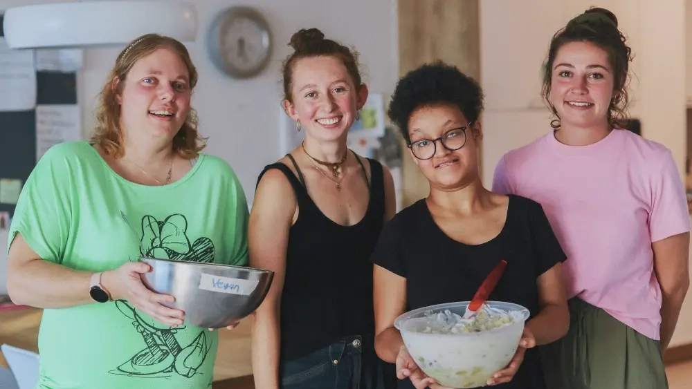 Vier junge Frauen in der Küche, zwei tragen Schüsseln in den Händen.