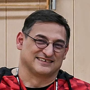 Sedat Oezbicerler: ein Mann mit kurzen dunklen Haaren und Brille. Er sitzt in Jeans und einer Sportjacke im Rollstuhl in einer Turnhalle.