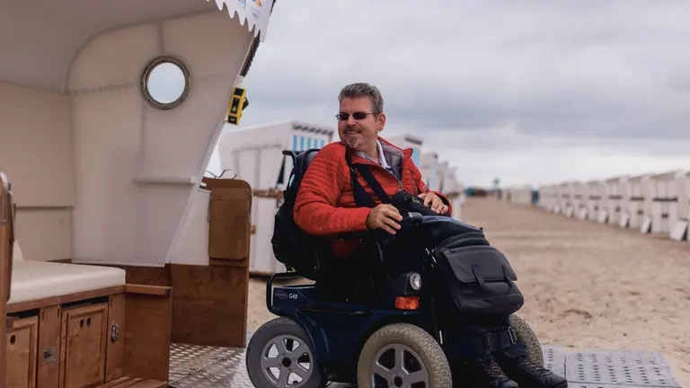 Am Strand fährt ein Rollstuhlfahrer aus einem Strandkorb.