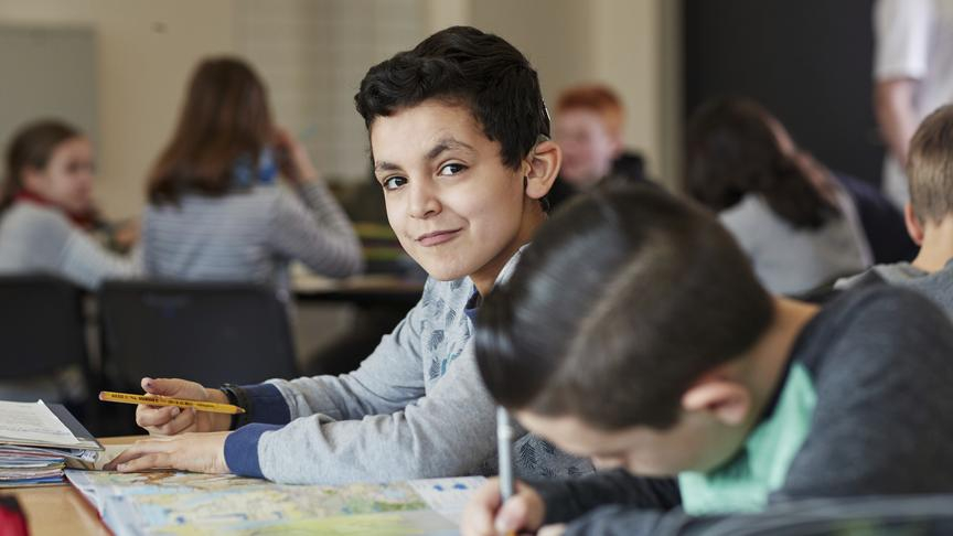 Ein Junge sitzt in einem vollen Klassenzimmer. Er hält einen Stift in der Hand und schaut in die Kamera.