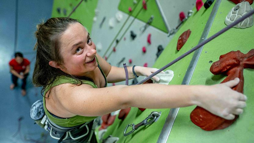 Eine junge Frau klettert an einer Wand in einer Kletterhalle. Sie wird von einem Mann an einem Seil gesichert.