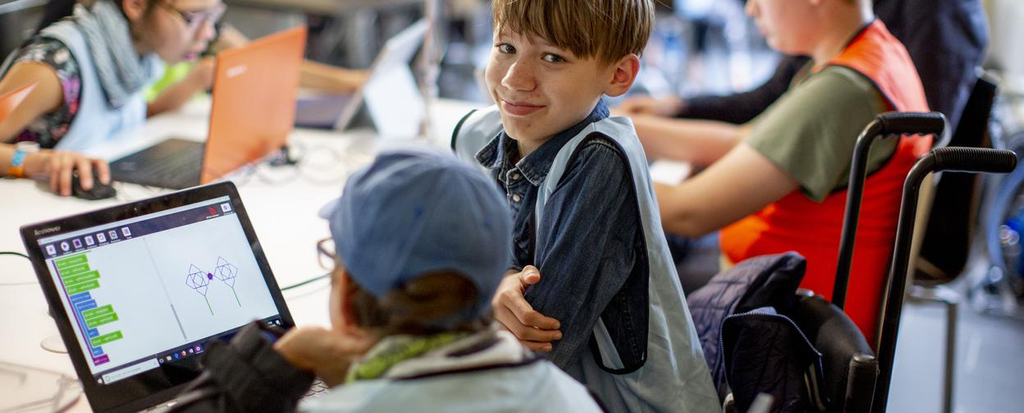 Zwei Jungen sitzen in einem Klassenzimmer gemeinsam vor einem Laptop. Einer der Jungen sitzt im Rollstuhl.