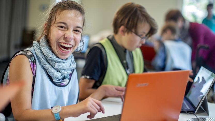 Ein Mädchen sitzt an einem Laptop und lächelt in die Kamera. Neben ihr sitzt ein Junge an einem Laptop. 