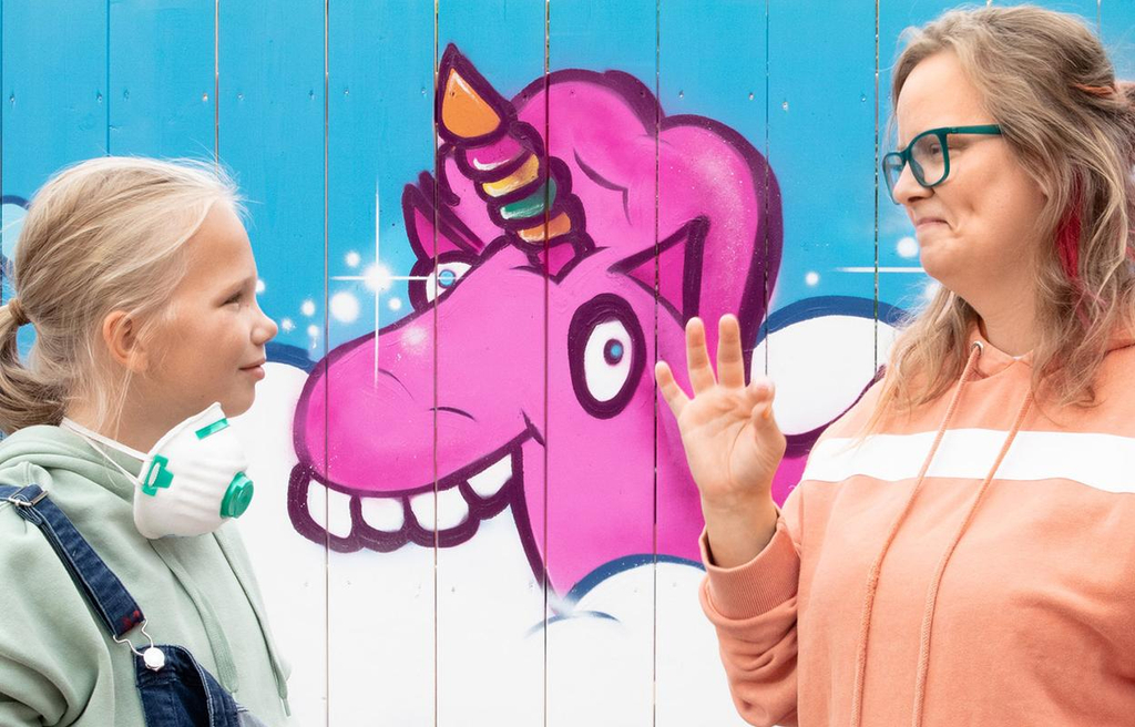 Die gehörlose Sylvia steht mit ihrer Tochter Lea vor einer Wand, auf der ein buntes Graffiti von einem Einhorn zu sehen ist.