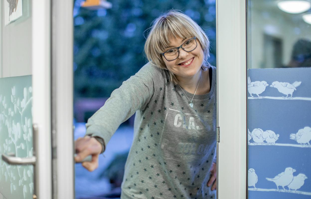 Eine junde Frau öffnet eine Tür und lächelt in die Kamera