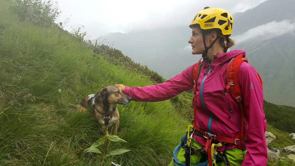 Eine junge Frau mit Helm steichelt einen Hund in einer Berglandschaft 