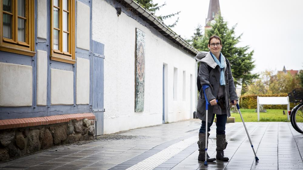 Agnes steht mit ihren Krücken auf einem Fußweg. Im Hintergrund ist ein Kirchturm zu sehen.